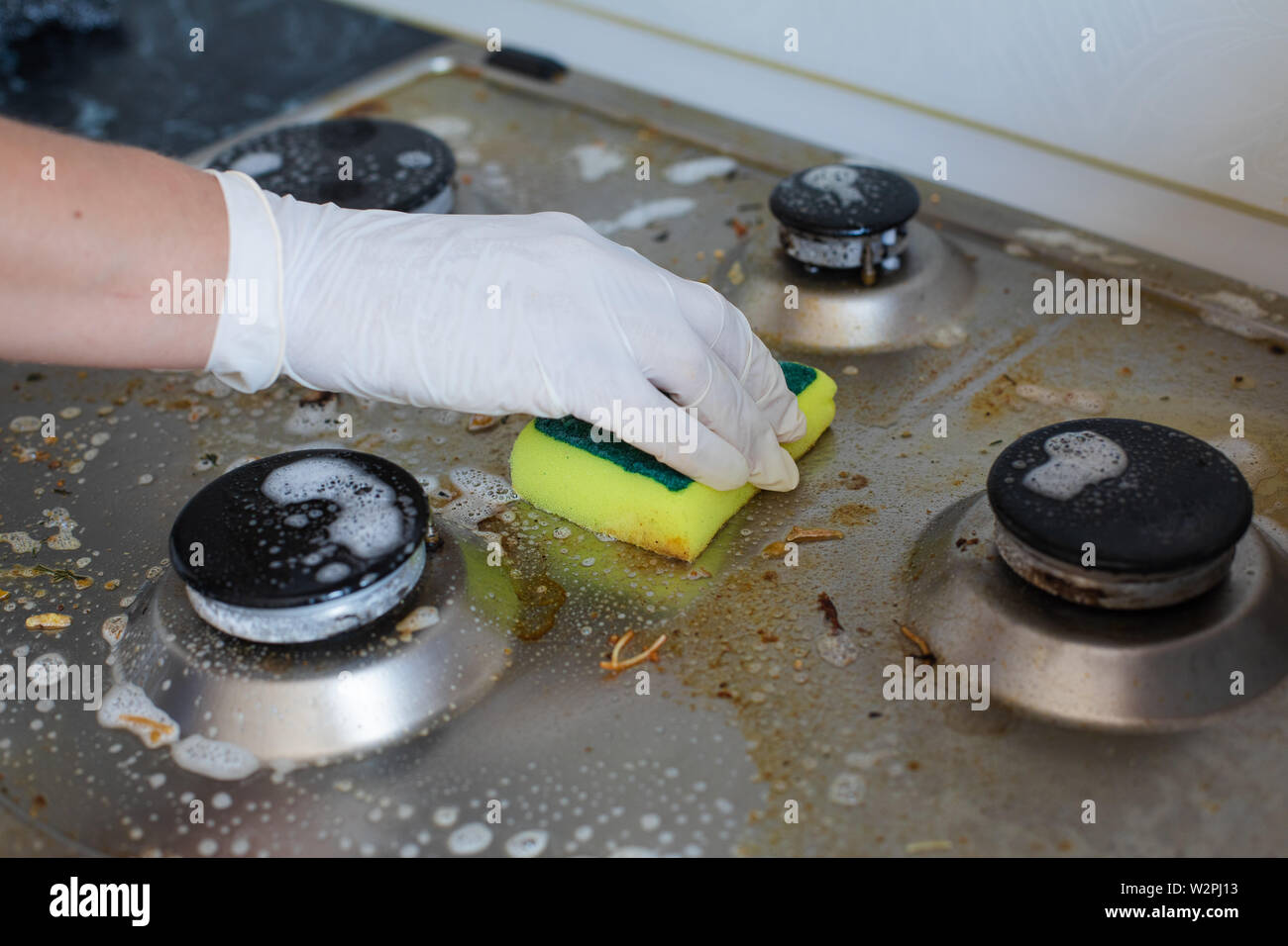 Domestic female hand wearing gloves cleaning dirty stove after cooking using sponge for washing