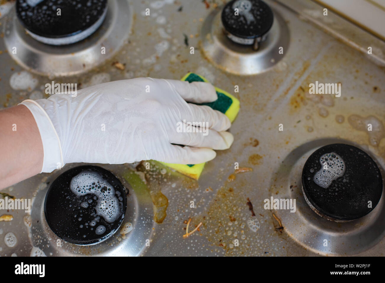 Domestic female hand wearing gloves cleaning dirty stove after cooking using sponge for washing