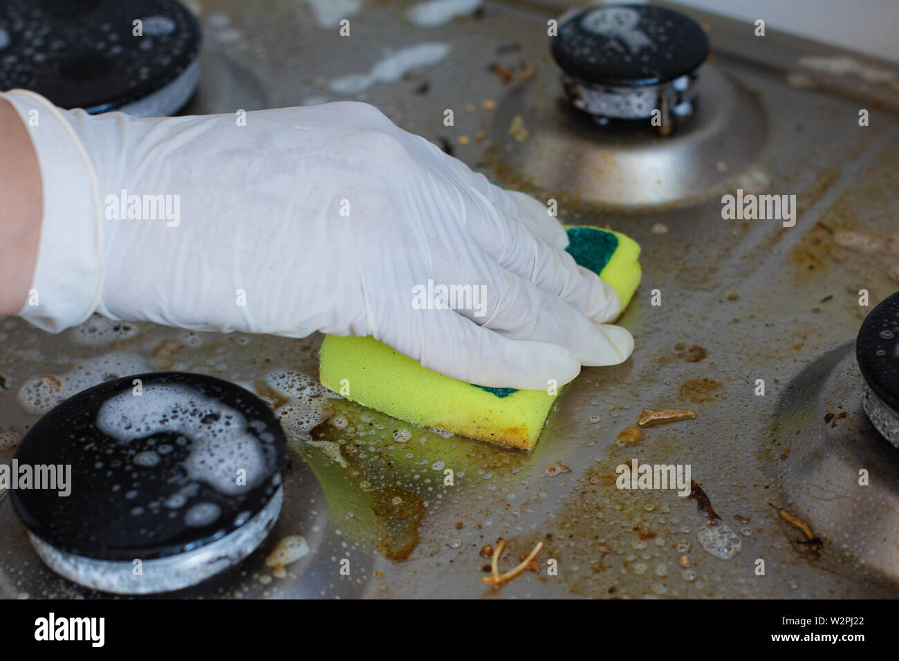 Domestic female hand wearing gloves cleaning dirty stove after cooking using sponge for washing