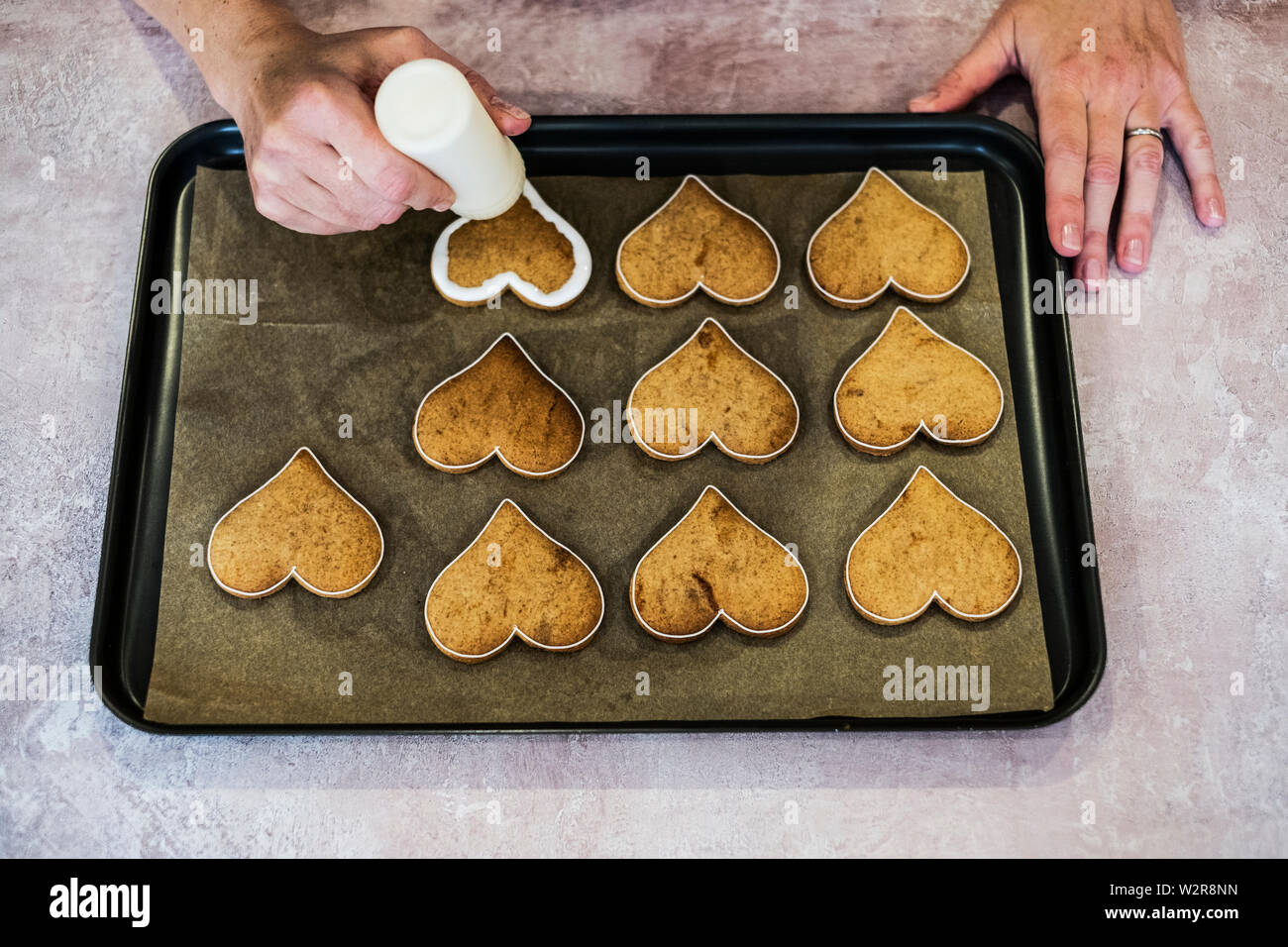 High angle close up of person decorating freshly baked hart-shaped cookies with piped white icing, flood icing technique. Stock Photo