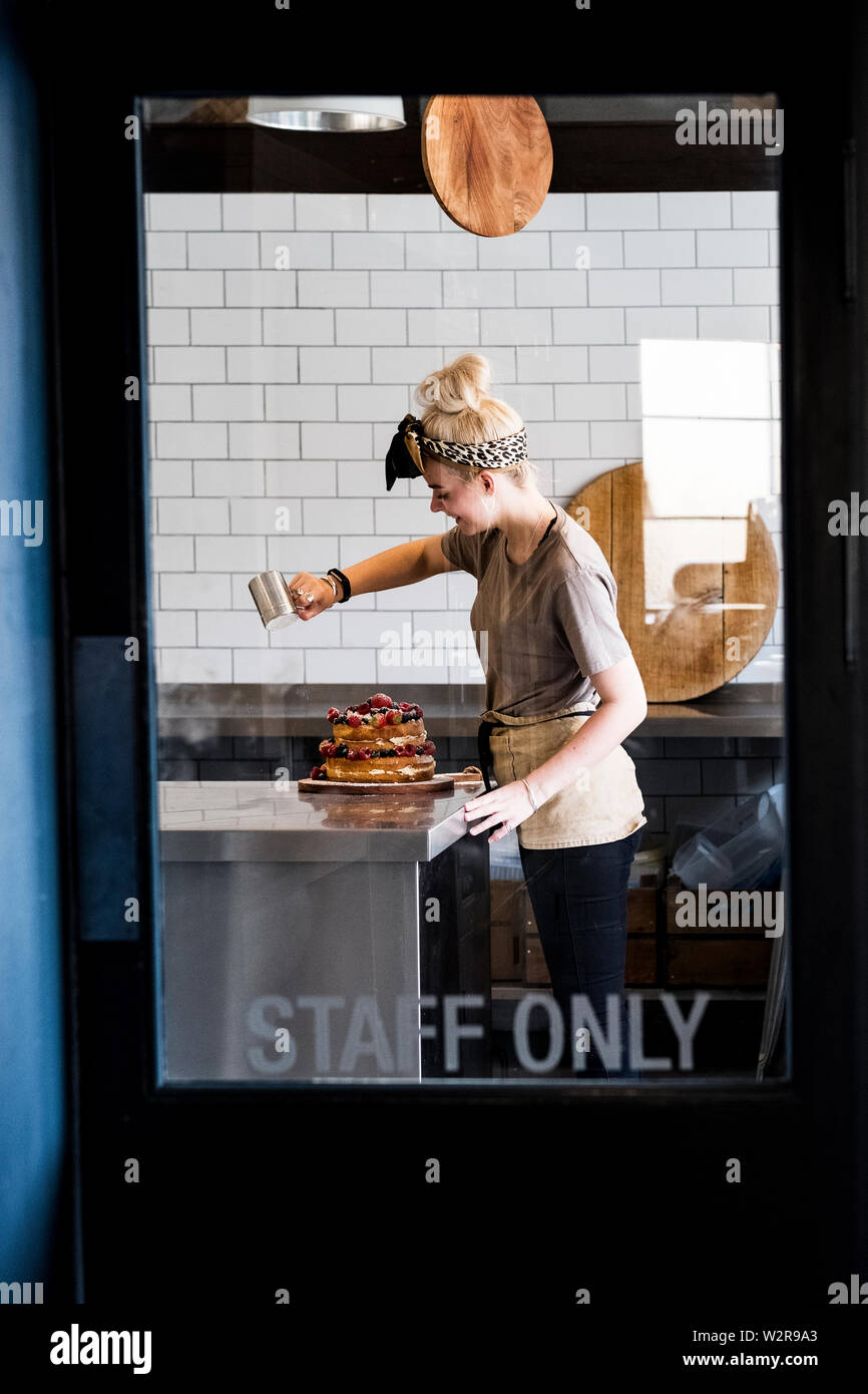 View through a door marked Staff Only, of a cook working in a commercial kitchen sprinking icing sugar over a layered cake with fresh fruit. Stock Photo