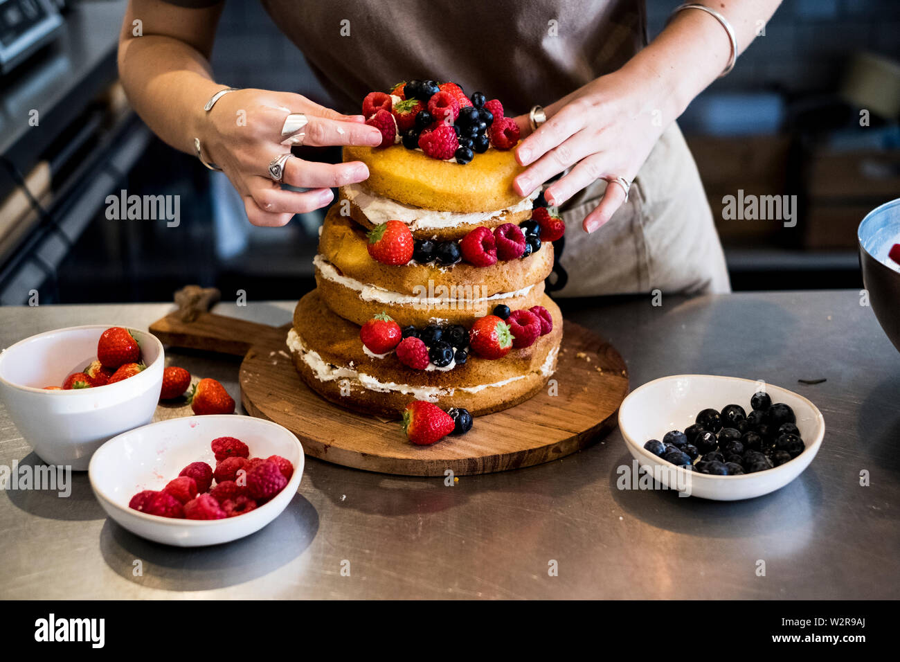a cook working in a commercial kitchen arranging fresh fruit over a layered cake with fresh cream. Stock Photo
