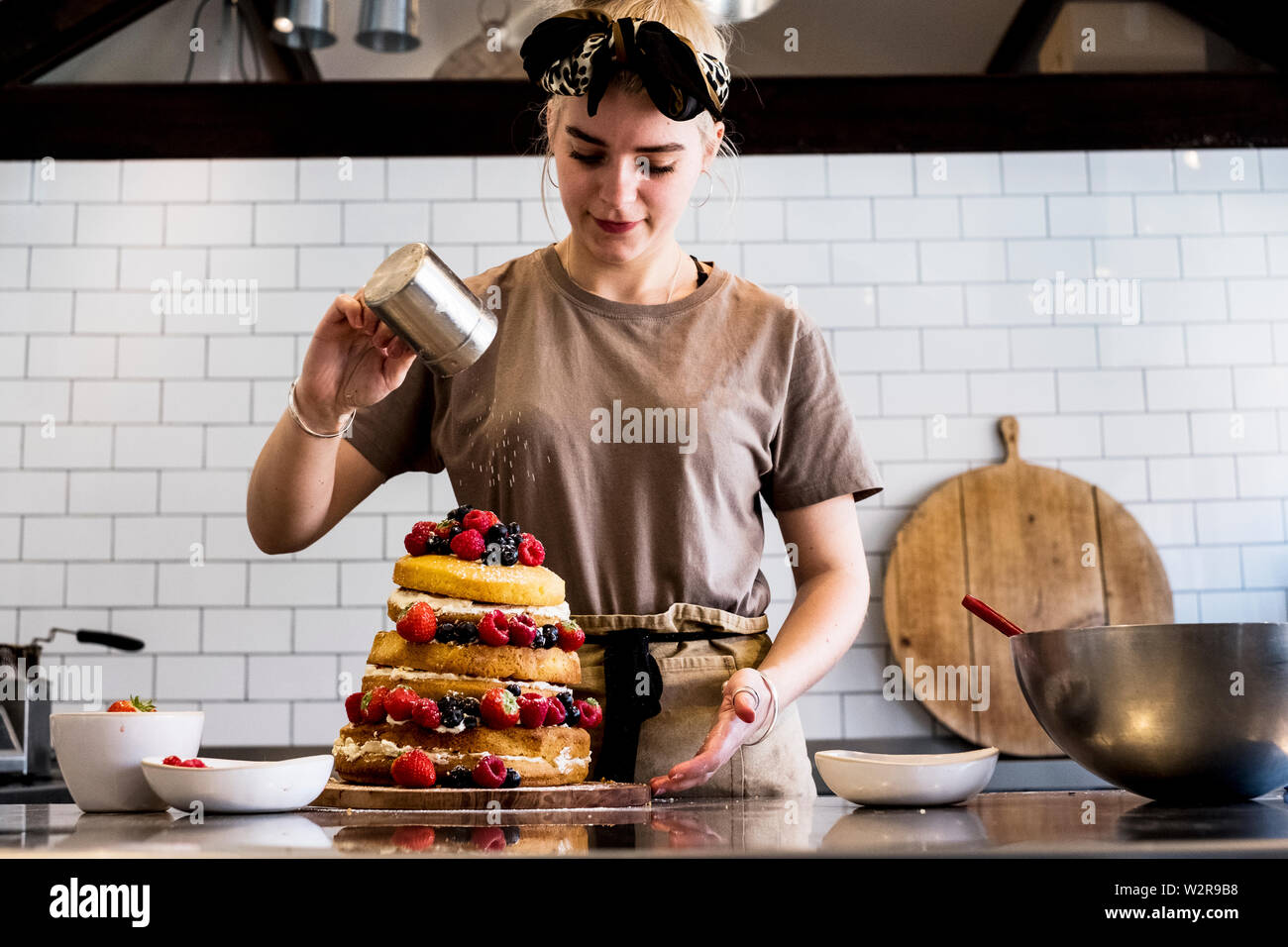 A cook working in a commercial kitchen sprinking icing sugar over a layered cake with fresh fruit. Stock Photo