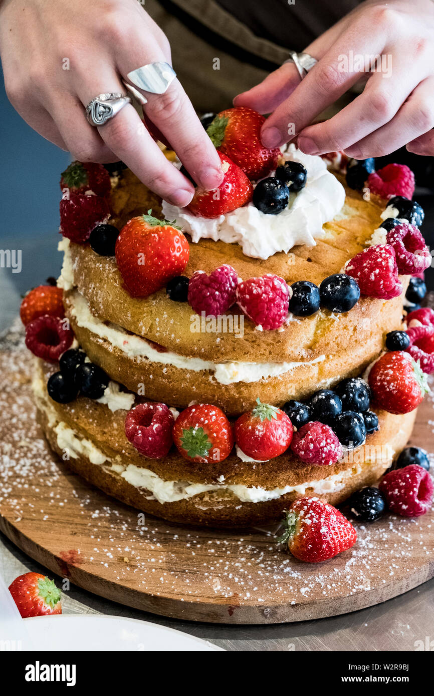 A woman cook asembling a layer cake with fresh cream and fresh fruit, strawberries and blueberries. Stock Photo