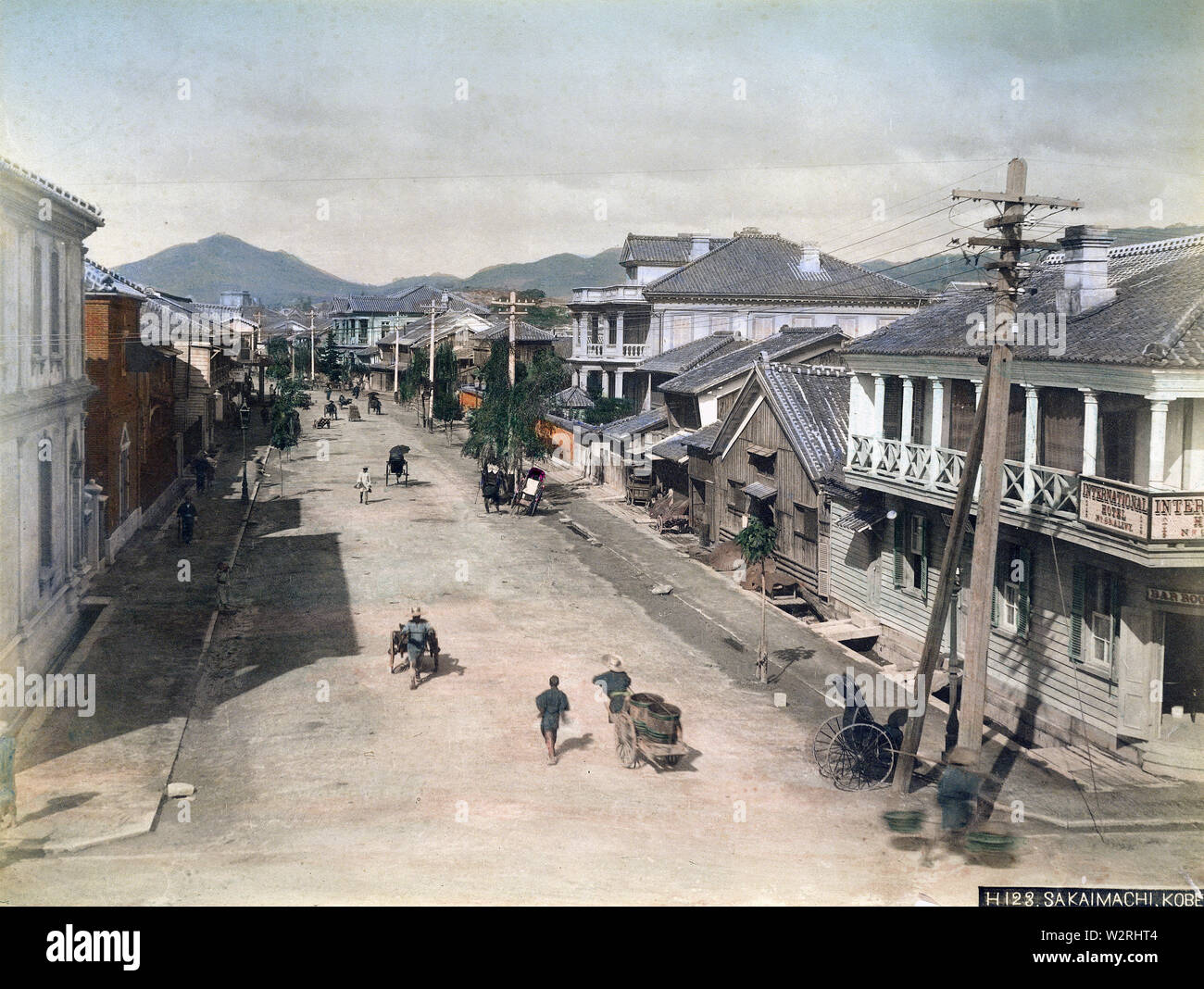 [ 1890s Japan - Main Street in Kobe ] —   A rickshaw puller rests in his carriage, while vendors go about their business, on Sakaemachi in Kobe, Hyogo Prefecture. The building on the right corner is the International Hotel.  19th century vintage albumen photograph. Stock Photo