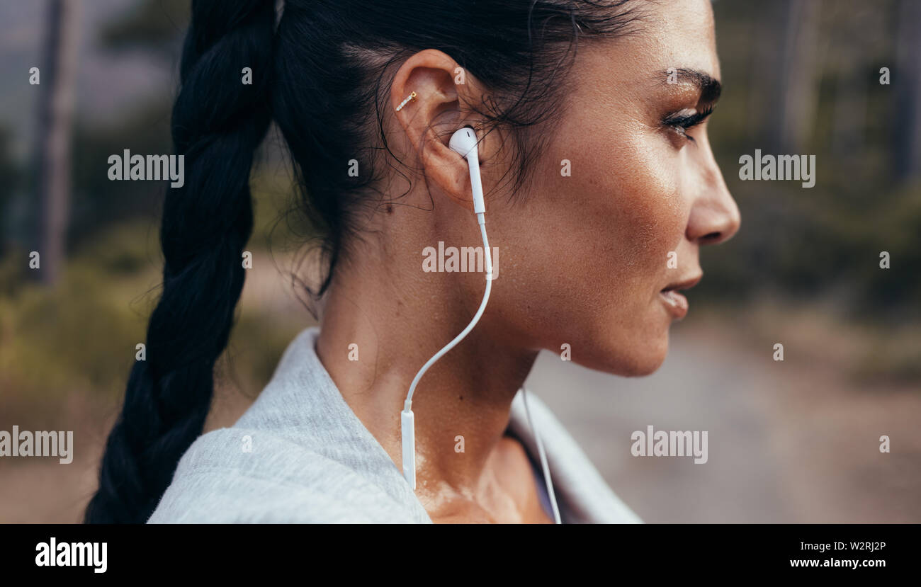 Side view of confident young woman with earphones standing outdoors after a morning run. Female athlete taking a rest after outdoor workout. Stock Photo