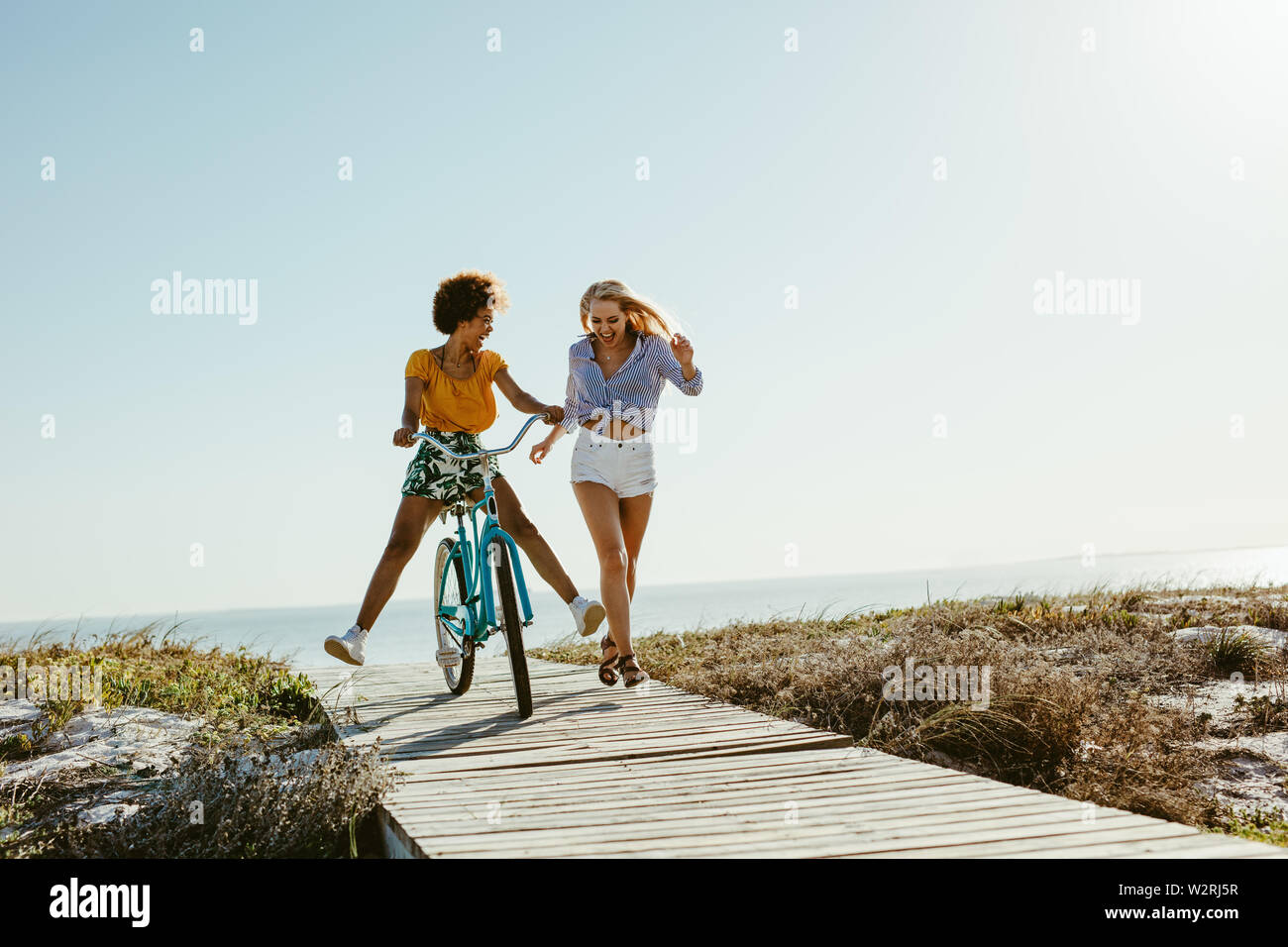 Two young women having fun with a bicycle at the beach. Woman running with friend riding a bike on boardwalk. Stock Photo