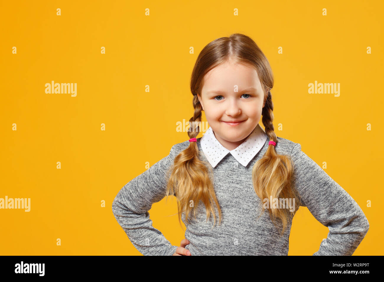 Portrait of a cute little girl on a yellow background. Child schoolgirl made hands to the side and looks into the camera. The concept of education. Stock Photo