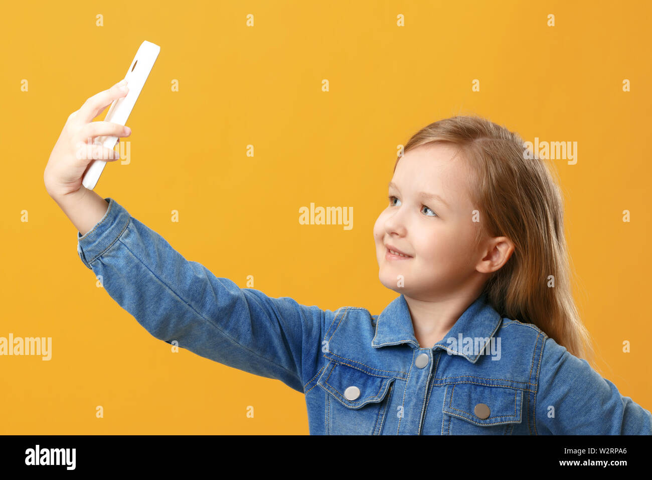 Portrait of a cute little girl in a denim shirt on a yellow background. The child holds the phone and takes a selfie Stock Photo