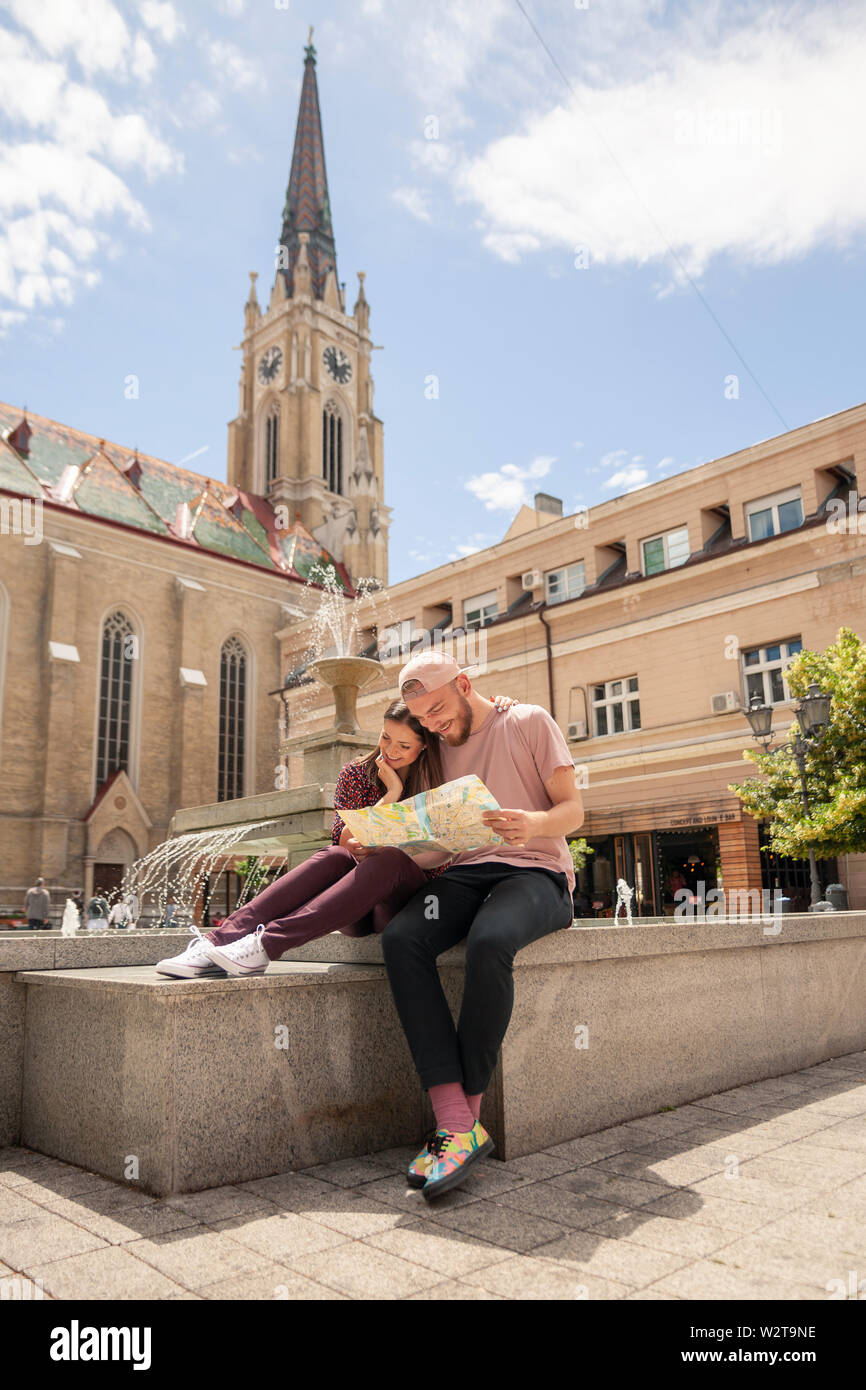 young couple sitting looking at a city map. In an old city European style square. Location: Serbia, Novi Sad. Stock Photo