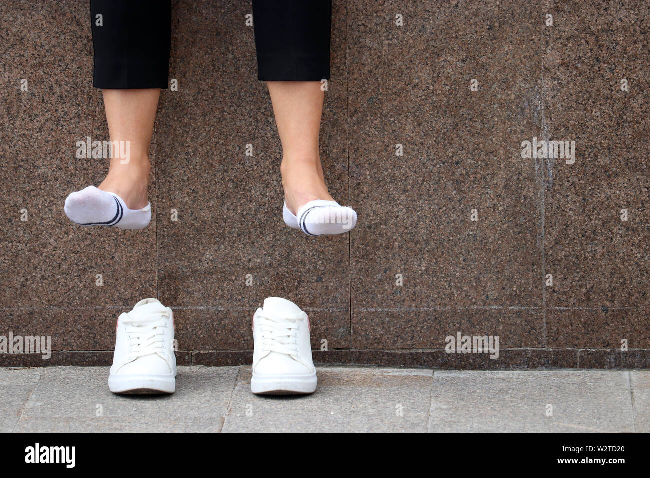 Tired legs, foot pain, barefoot woman with removed white sneakers resting on a street. Female legs in white socks dangling over shoes Stock Photo