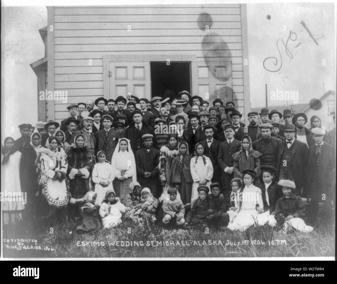 Eskimo wedding, St. Michael, Alaska. July 1st, 1906, 10 P.M. Photograph shows a demure bride, and solemn groom, standing slightly off-center and overshadowed by large group of wedding guests surrounding them outside building. Stock Photo