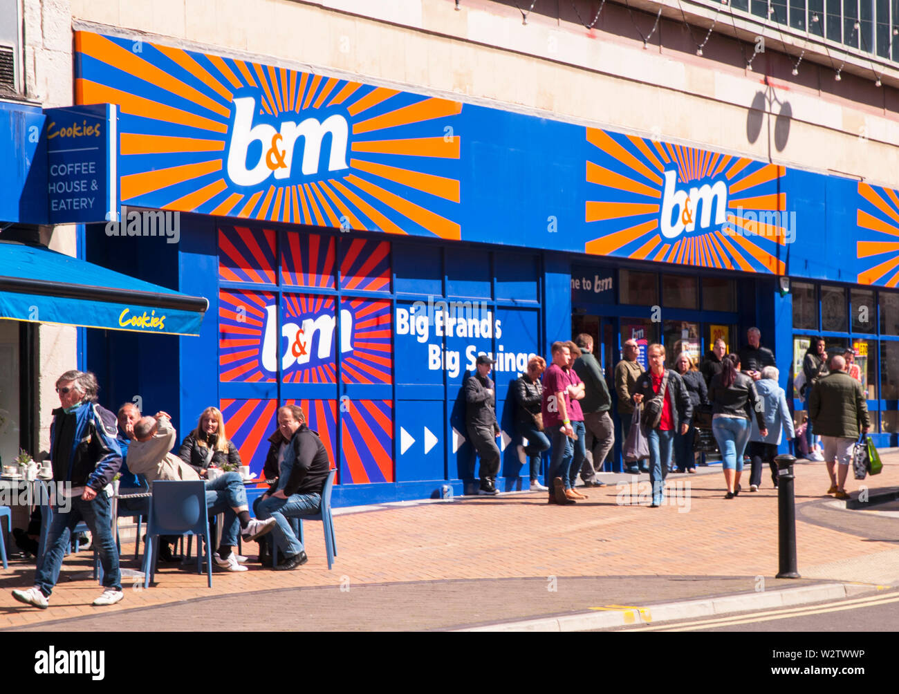 Men standing outside B&M Bargain discount store possible waiting for wifes / girlfriends to finish shopping  People also sitting outside cafe in sun Stock Photo