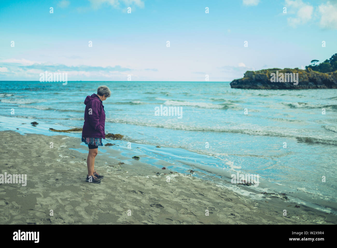 A senior woman is standing by the water's edge on the beach Stock Photo