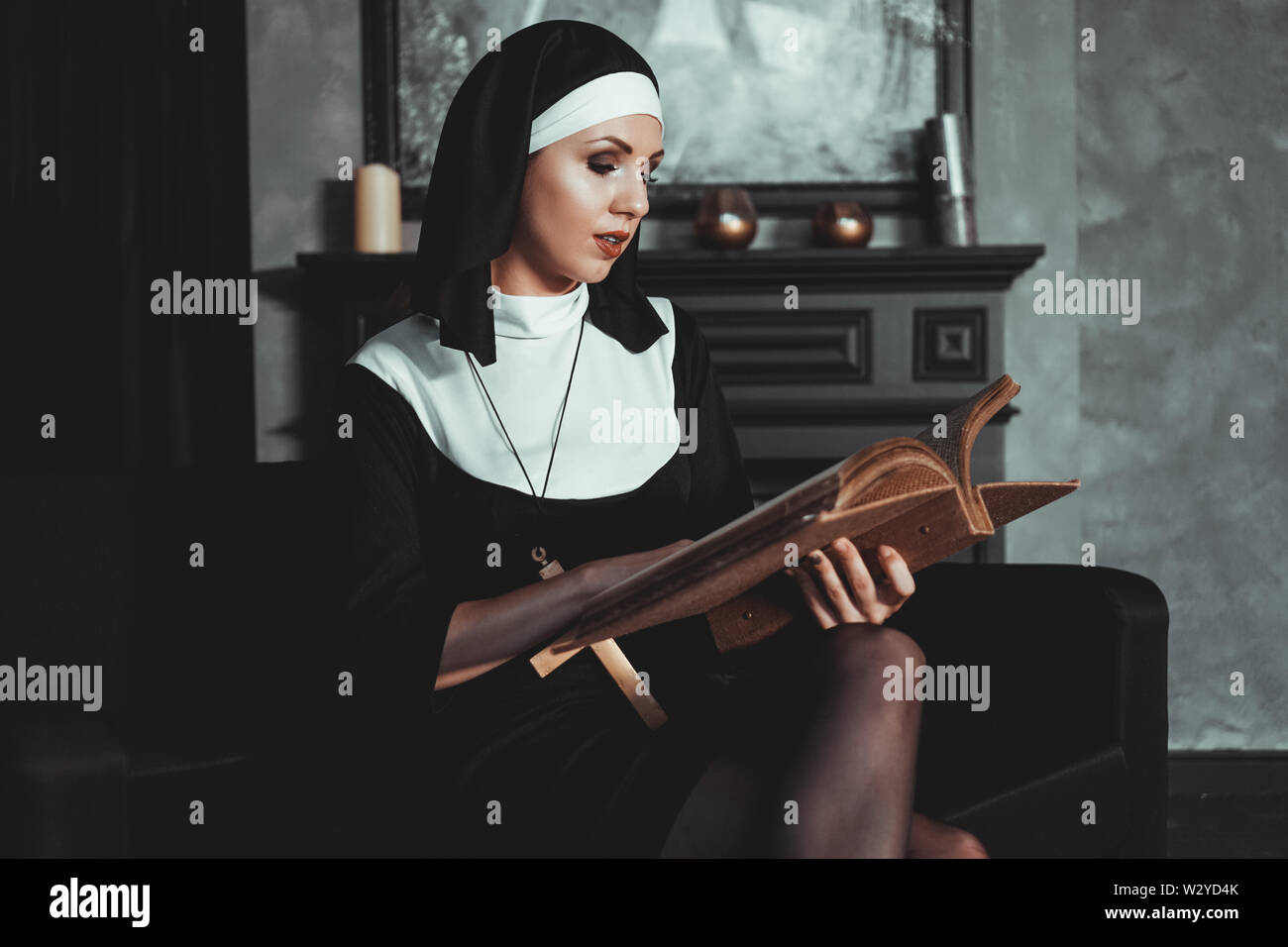 Beautiful young nun in religion black suit holds Bible and posing on camera with big book on a black background. Religion concept. Stock Photo