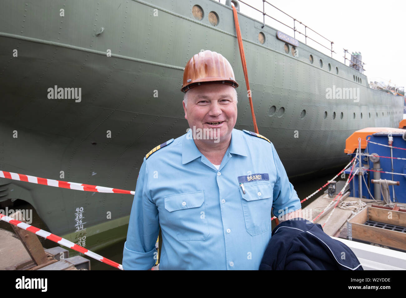 11 July 2019, Lower Saxony, Bern: Commander Nils Brandt exchanged his captain's cap for a helmet after the docking of the naval training ship 'Gorch Fock' at the Fassmer shipyard.  At the shipyard the 'Gorch Fock' is lifted out of the water. In an assembly hall, workers from the Elfleth shipyard are making further progress with the restoration of the ship. Photo: Jörg Sarbach/dpa Stock Photo