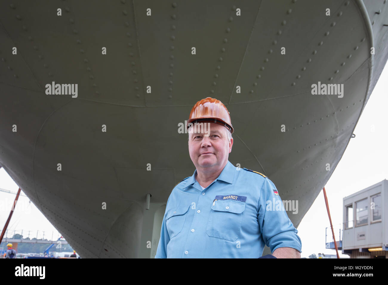 11 July 2019, Lower Saxony, Bern: Commander Nils Brandt exchanged his captain's cap for a helmet after the docking of the naval training ship 'Gorch Fock' at the Fassmer shipyard.  At the shipyard the 'Gorch Fock' is lifted out of the water. In an assembly hall, workers from the Elfleth shipyard are making further progress with the restoration of the ship. Photo: Jörg Sarbach/dpa Stock Photo