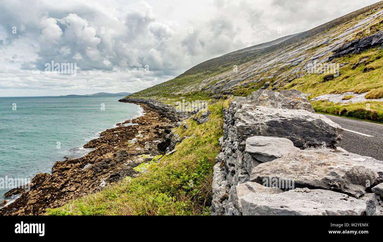 Beautiful Irish landscape with the sea and the rural coastal road along​​ the Burren, geosite and geopark, Wild Atlantic Way, cloudy spring day Stock Photo