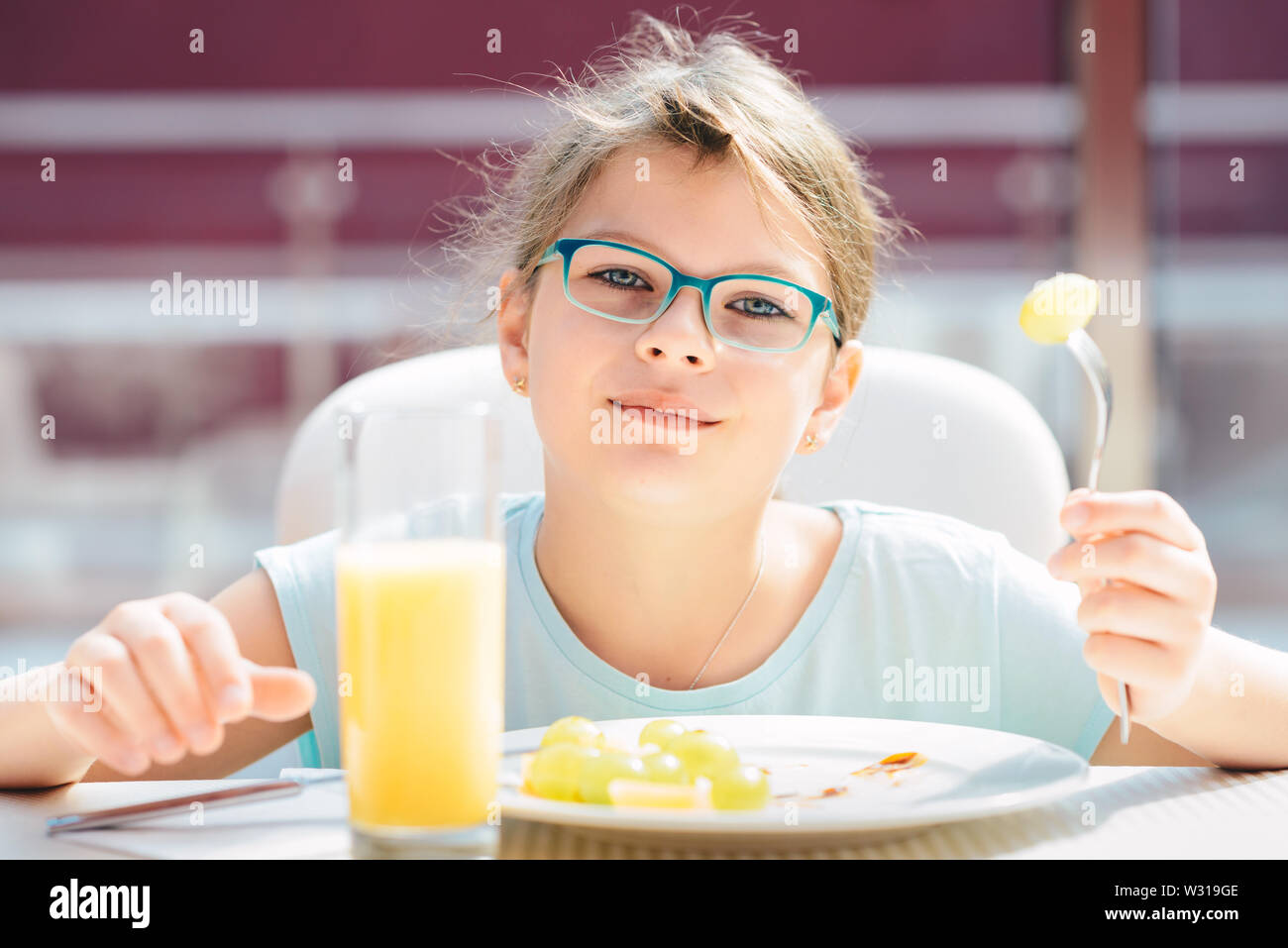Cheerful girl eating pancakes, fresh fruit and drinking orange juice