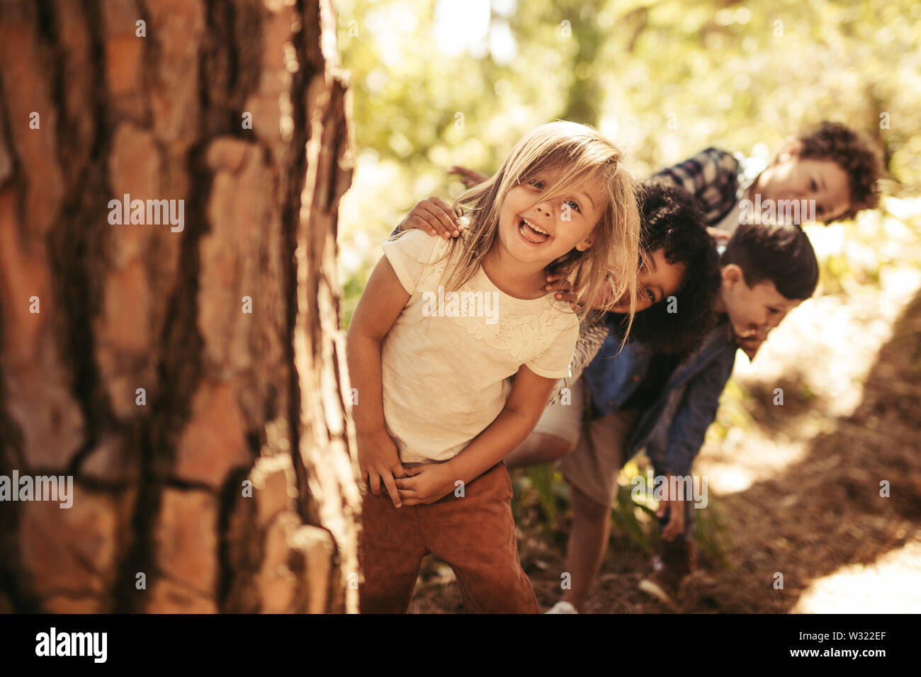 Group of kids standing in row and peeking behind a tree outdoors. Children playing hide and seek in a park. Stock Photo