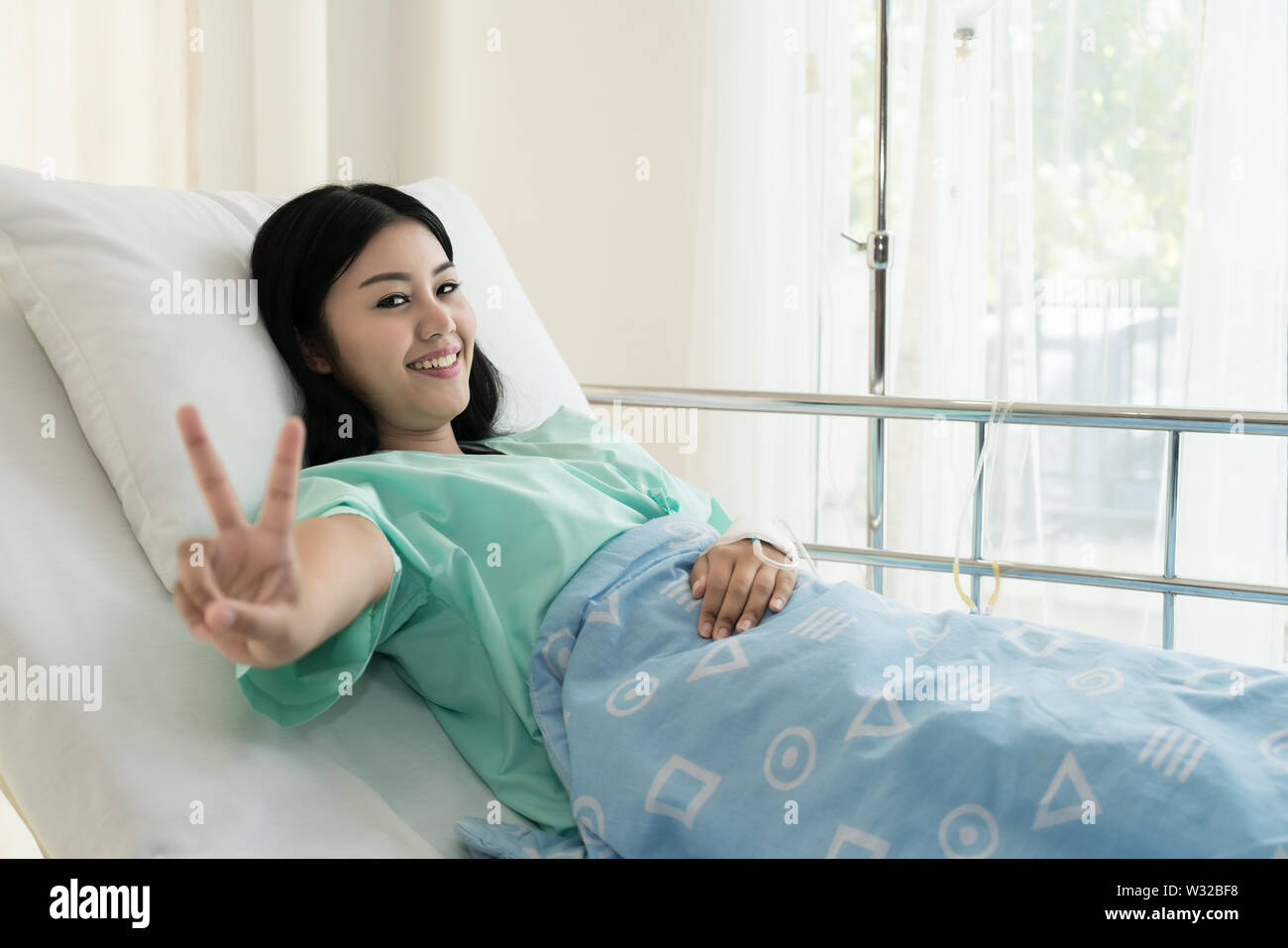 Young Asian patient woman lying at hospital bed with saline drip showing victory sign for cheerful. Drop of saline solution to help patient. Stock Photo
