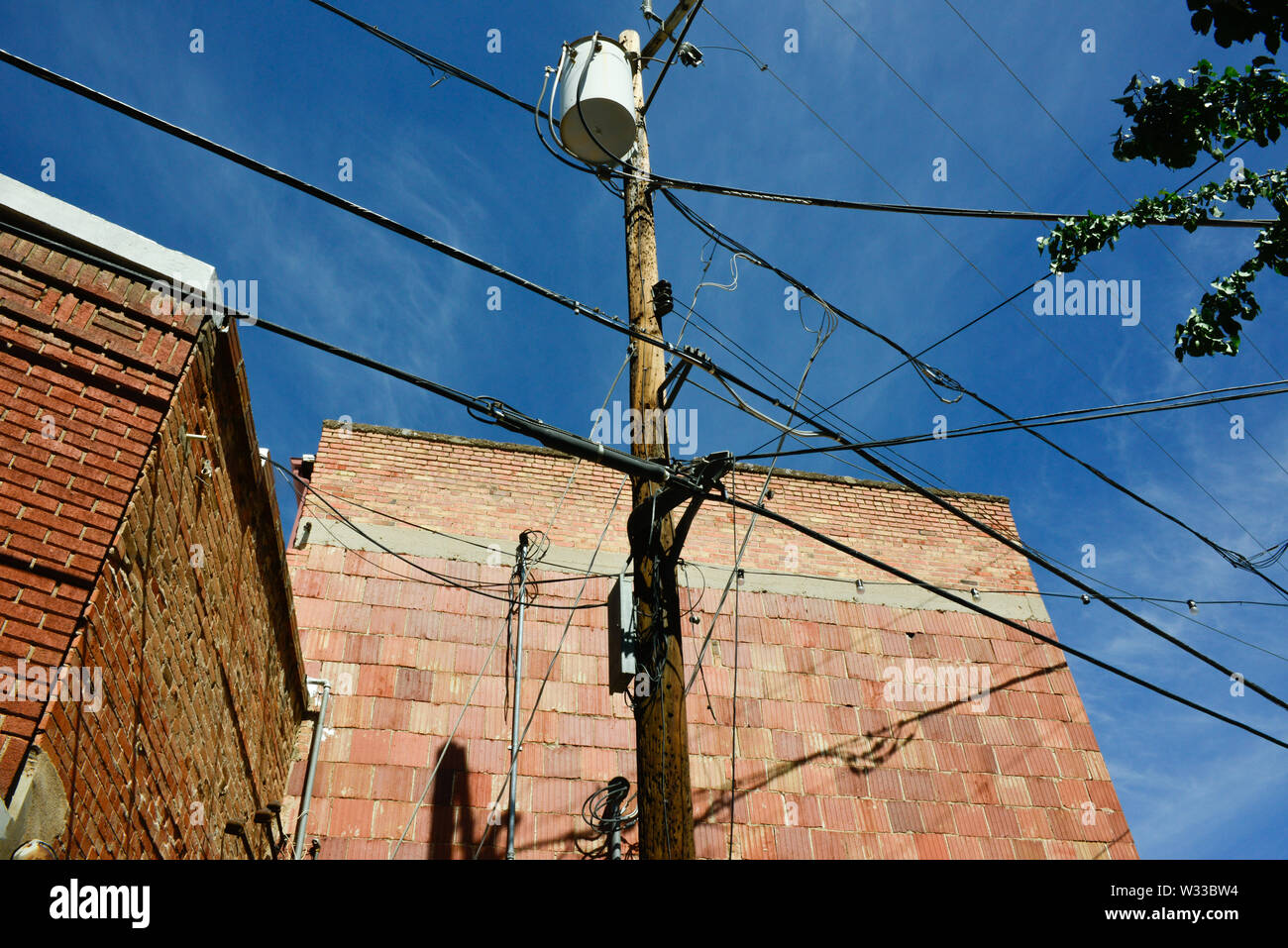 An upward view of several electrical lines and telephone cables intersecting at a wooden light pole against old red brick and concrete buildings, USA Stock Photo