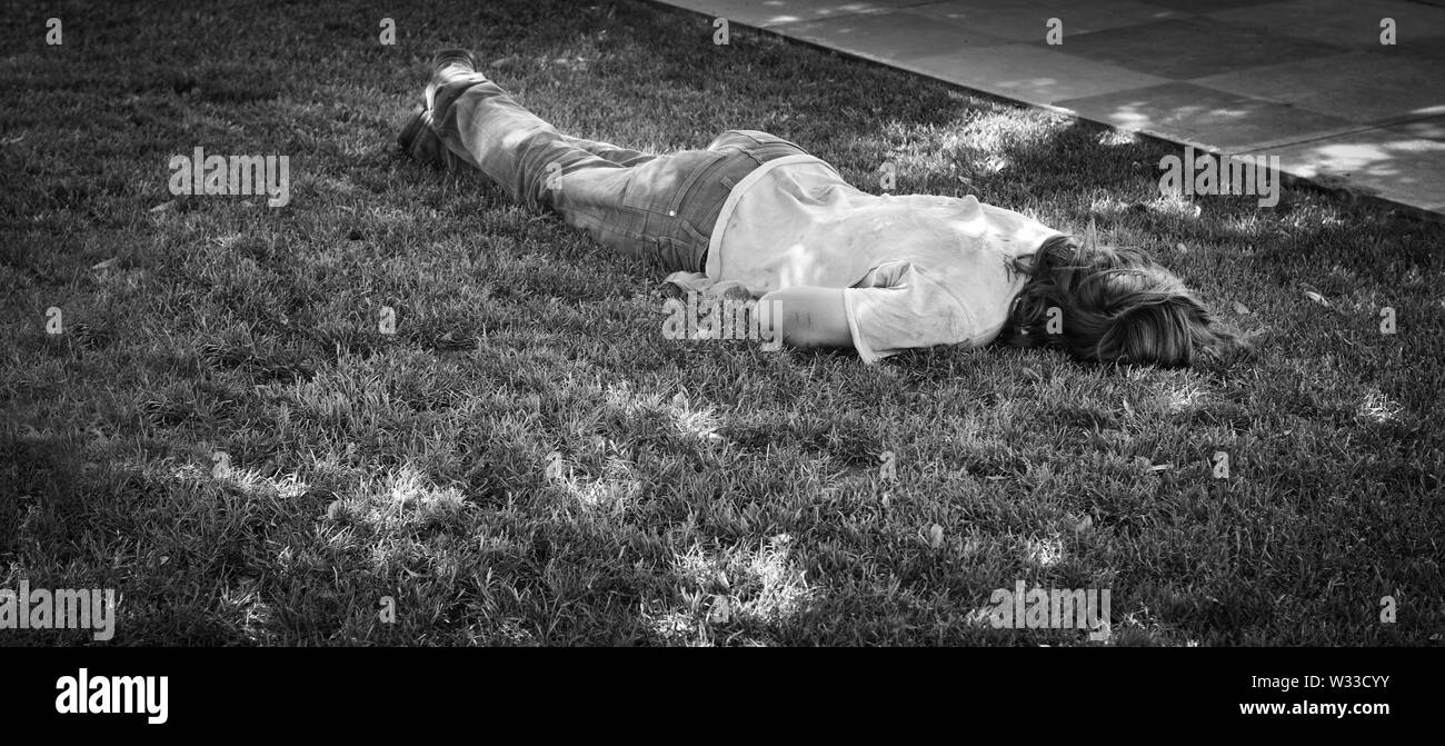 A Caucasion homeless man sleeps face down on the grass in a park in the summertime, in black and white Stock Photo