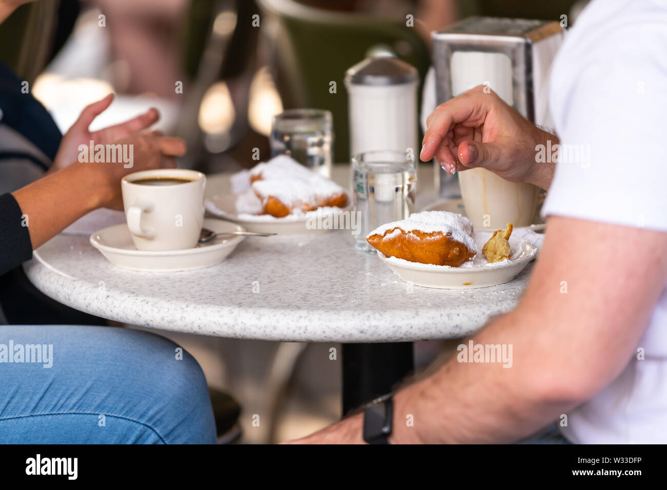 Closeup of couple sitting outside at sidewalk cafe by table drinking chicory coffee and eating deep fried beignet donut powdered with sugar Stock Photo