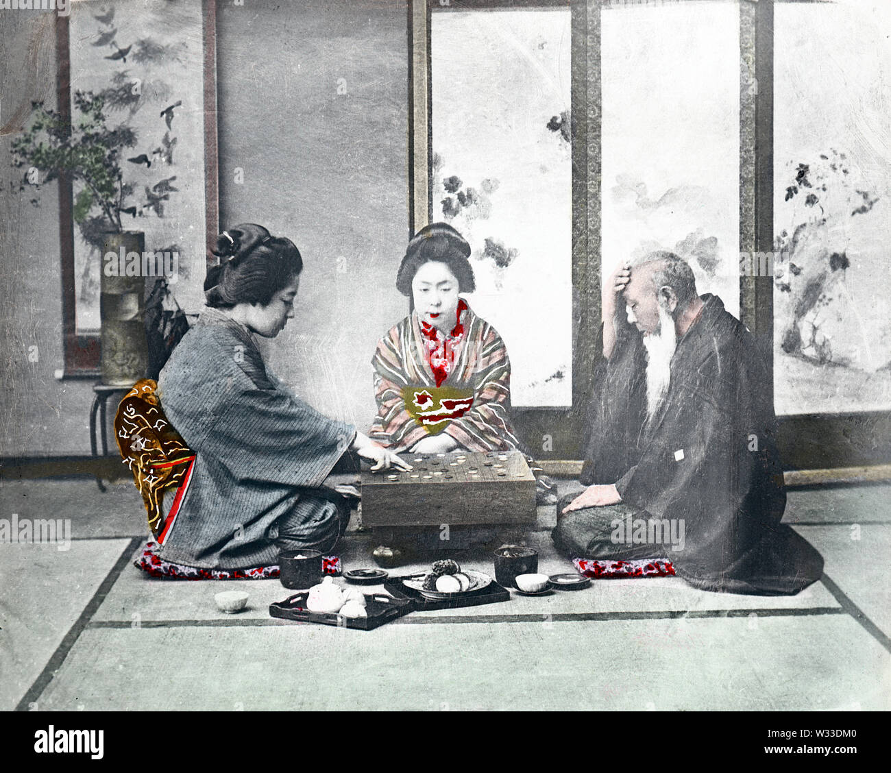 [ 1890s Japan - Playing Go ] —   Two young women in kimono and traditional hairstyles and an elderly man in a haori coat are seated around a board for the game of go.  The studio is arranged as a zashiki (guest room), with a screen, an ikebana flower arrangement and scroll in the back. A small tray with a teapot and two cups can be seen at the front.  19th century vintage glass slide. Stock Photo