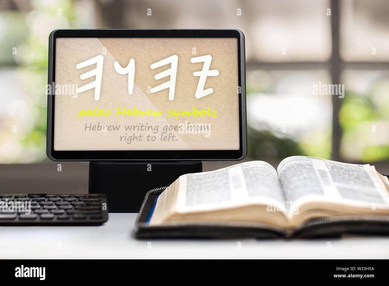 A tablet showing the paleo Hebrew symbols for the name of God on a desk with a keyboard, opened holy bible and cross in background. Stock Photo