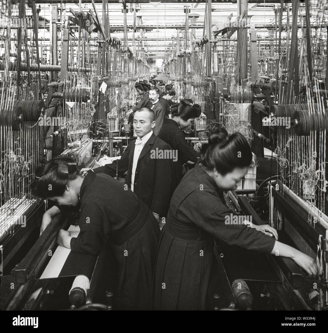 [ 1900s Japan - Japanese Silk Factory ] —   A man in suit looks on as women in uniform are weaving silk at the Kiryu Orimono KK (桐生織物株式会社, Kiryu Textile Company Ltd.), a Japanese silk factory in Kiryu, Gunma Prefecture. The company was launched as the Nihon Orimono KK (日本織物株式会社, Japan Textile Company, Ltd.) in 1887 (Meiji 20). At the time it was Japan’s largest and most modern silk factory where the complete production process was done by machinery.  20th century vintage glass slide. Stock Photo
