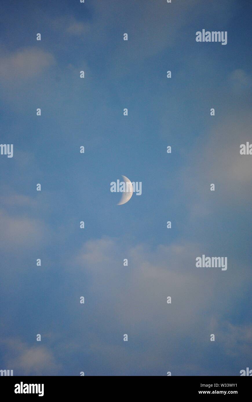 Crescent moon in the daytime with white clouds  and a blue sky Stock Photo