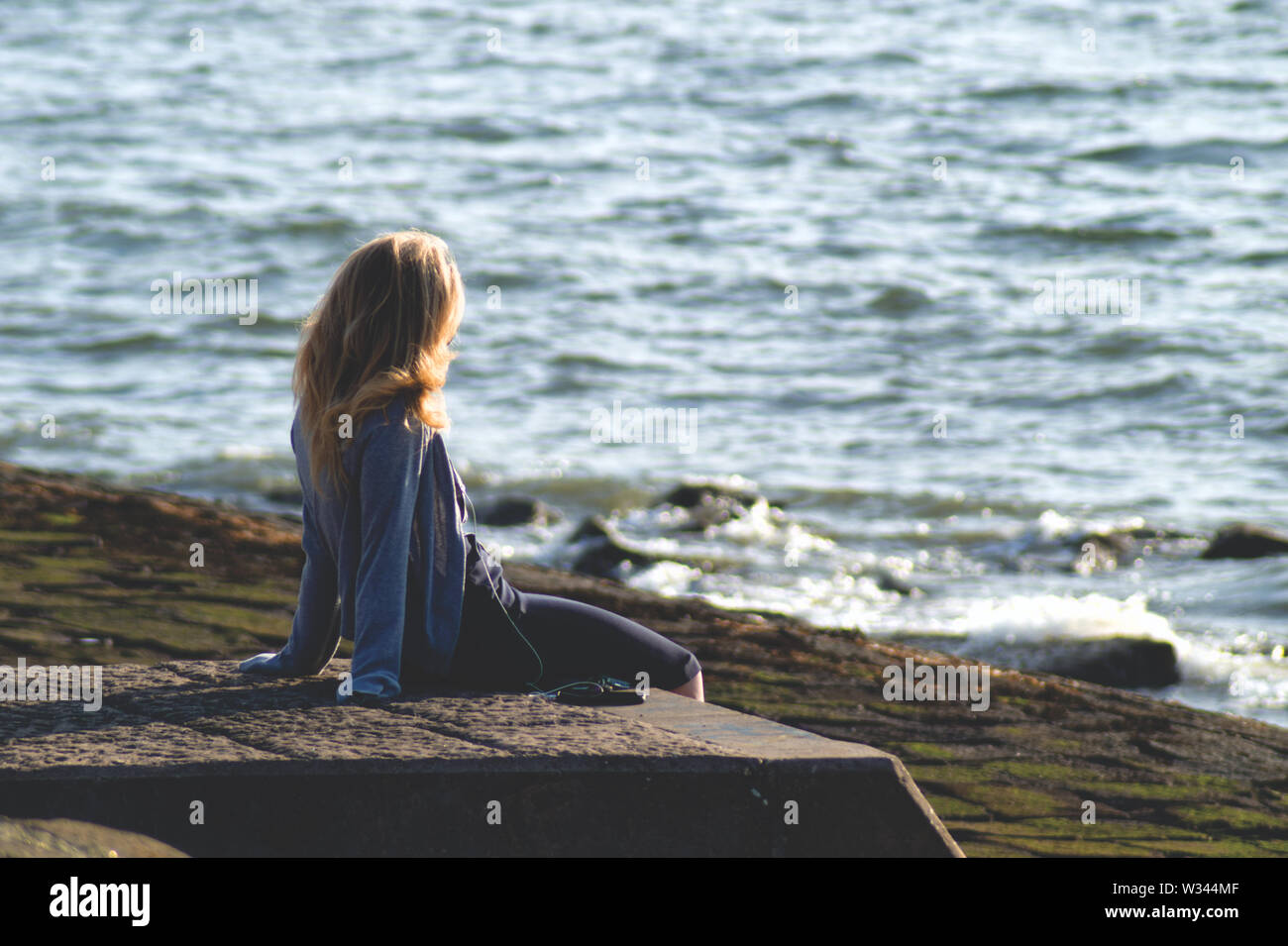 A single blond woman sitting on a stone pier looking out at the sea listening to music Stock Photo