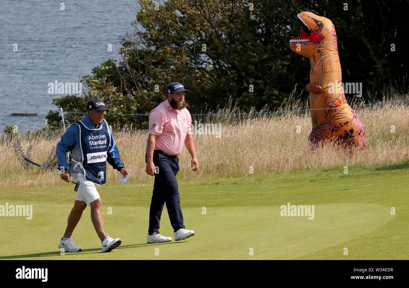 England's Andrew Johnston on the 4th green as a spectator in a Dinosaur costume walks by during day two of the Aberdeen Standard Investments Scottish Open at The Renaissance Club, North Berwick. Stock Photo