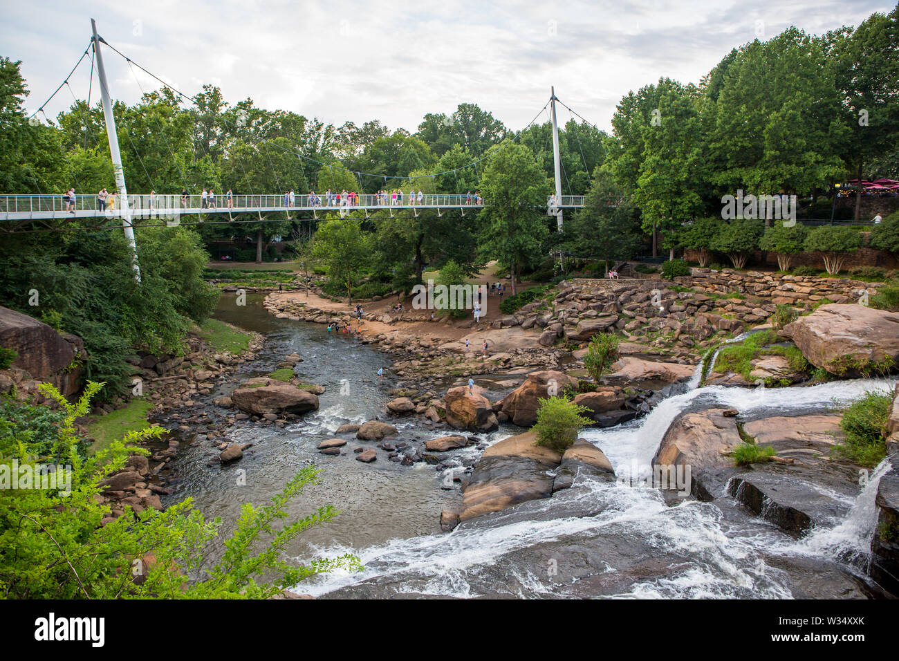 Visitors to Falls Park linger on Liberty Bridge overlooking the Reedy River waterfalls in downtown Greenville, South Carolina. Stock Photo