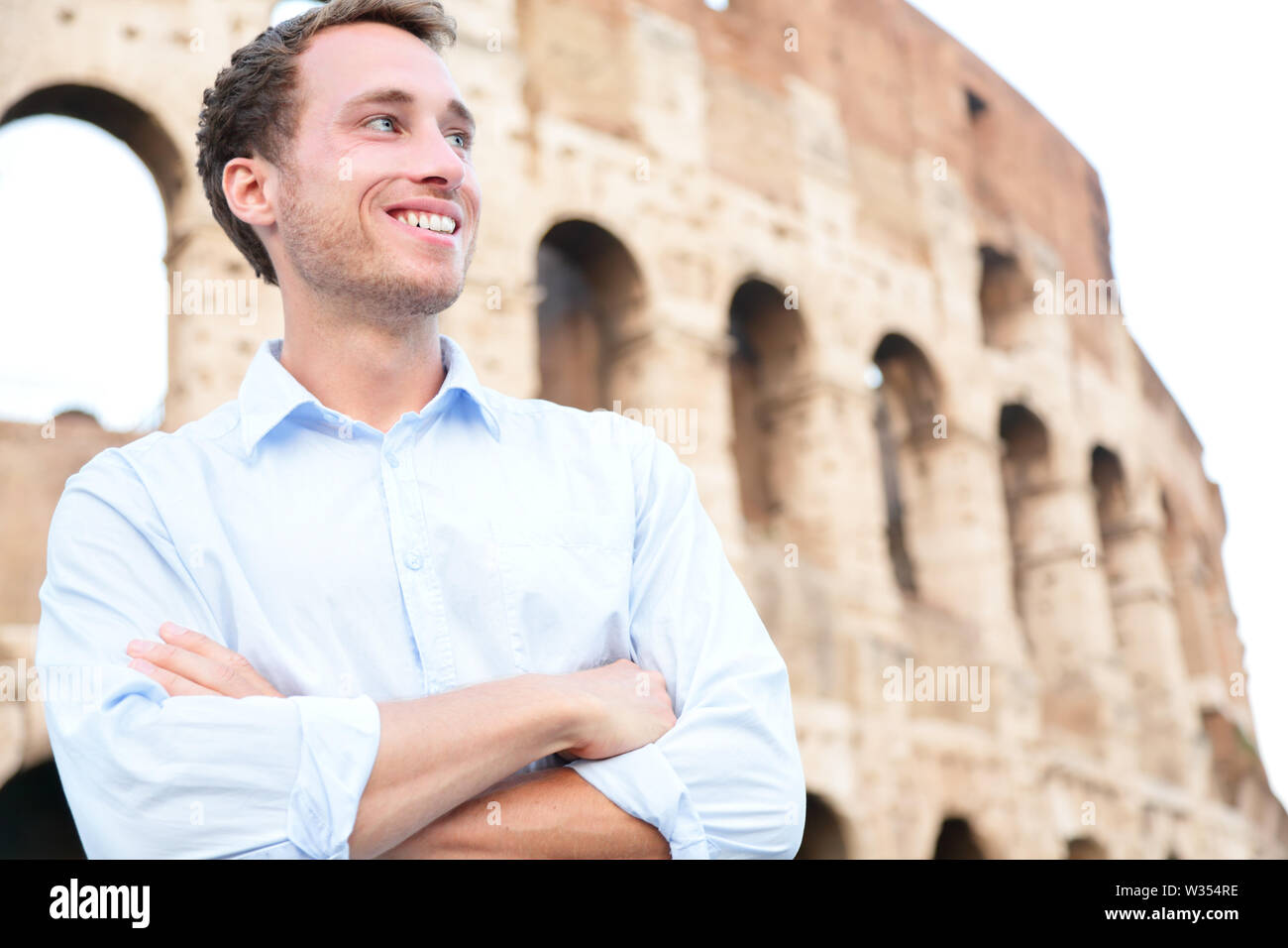 Young casual business man portrait by Colosseum, Rome, Italy. Proud confident happy smiling cross-armed businessman in shirt standing outdoor looking to side. Male Caucasian man in his 20s. Stock Photo
