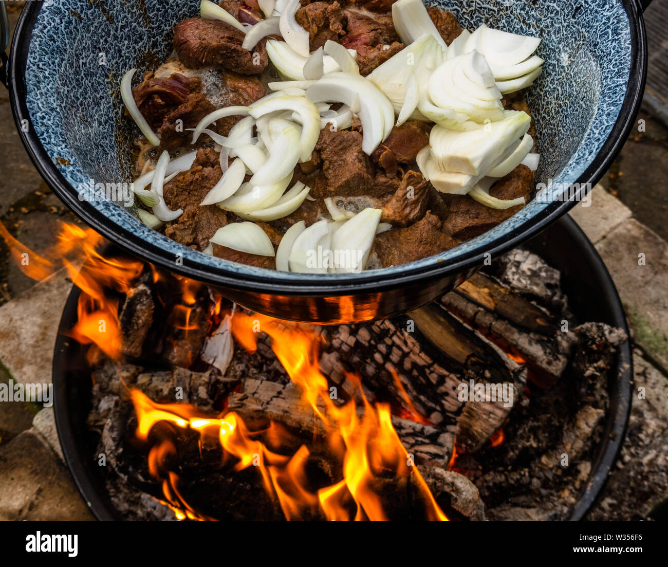 White onions and roasted pieces of beef hang in a pot over an open wood fire for the preparation of kettle goulash. Stock Photo