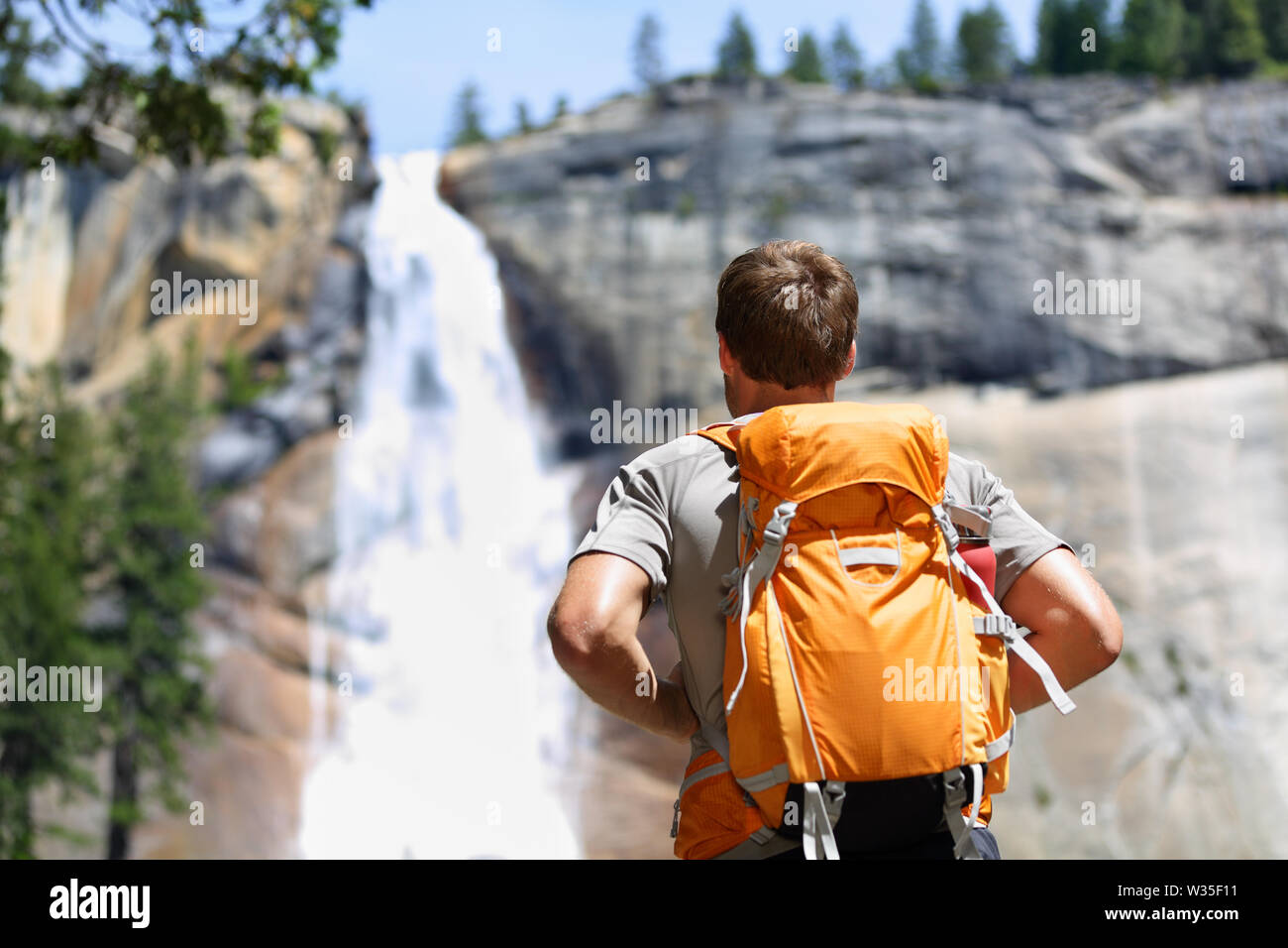 Hiker hiking with backpack looking at waterfall in Yosemite park in beautiful summer nature landscape. Portrait of male adult back standing outdoor. Stock Photo