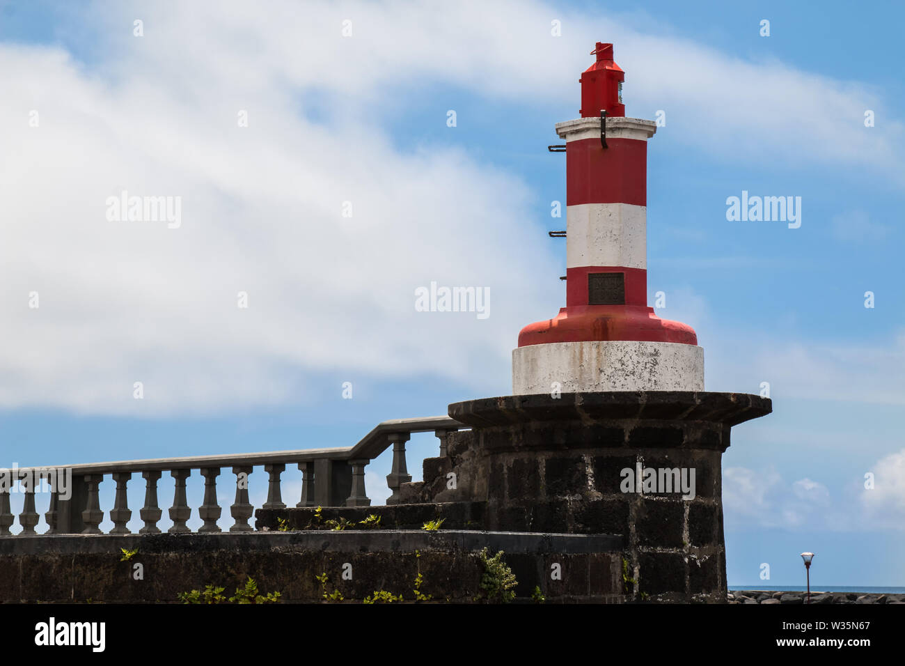 Stone wall and fence on the promenade along the Atlantic ocean. On the corner small red-white lighthouse. Blue sky with white clouds. Povoacao, Sao Mi Stock Photo