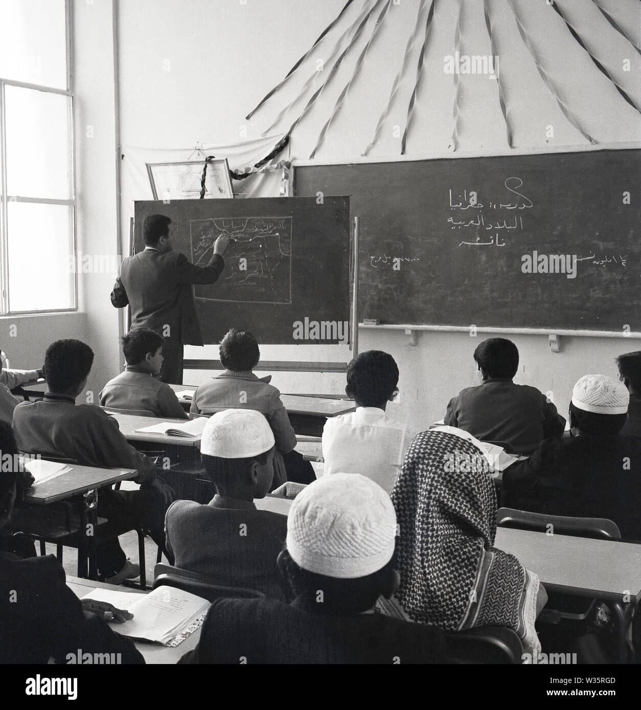 1960s, historical, arab schoolboys stting in a classroom, with a male teacher at one of the two blackboards in the room, drawing with chalk a diagram. Stock Photo