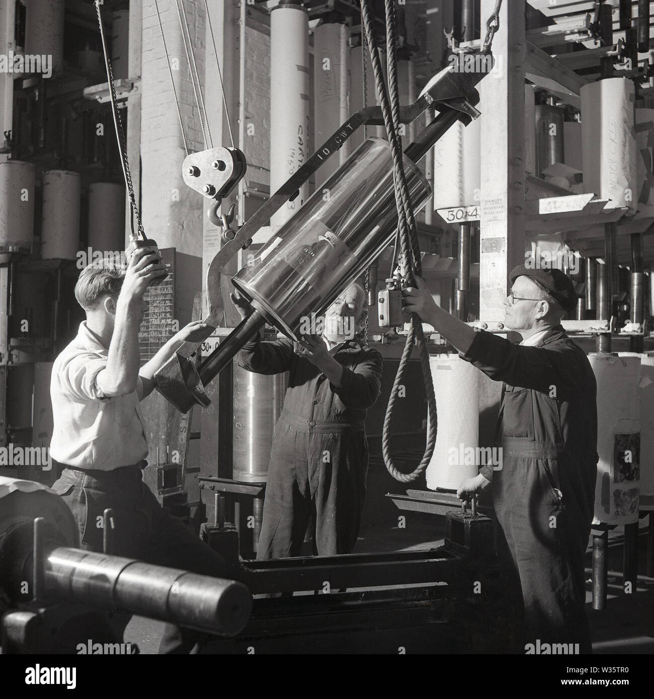 1950s, historical, male workers at the print works of Hazell, Watson & Viney holding a pulley as they move a stainless steel printing drum, England, UK. Based in Aylesbury, Buckinghamshire, the printing and publishing company, which started in business in 1839 and grew significantly after WW2. In the early 1960s, it merged with Purnell & Sons to form the British Printing Corporation (BPC), a company taken over by press baron Robert Maxwell in 1981. Stock Photo