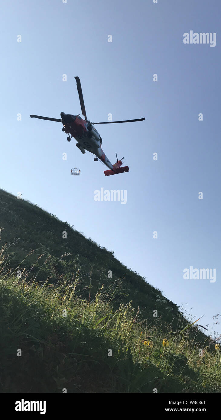 An MH-60 Jayhawk helicopter crew from Coast Guard Air Station Kodiak hoists an injured hiker from North Sister Mountain on Kodiak Island, Alaska, July 11, 2019. The 55-year-old woman reportedly fractured her leg while hiking and was unable to traverse the steep terrain back down the mountain. U.S. Coast Guard photo by Petty Officer 1st Class Nadia Laub. Stock Photo