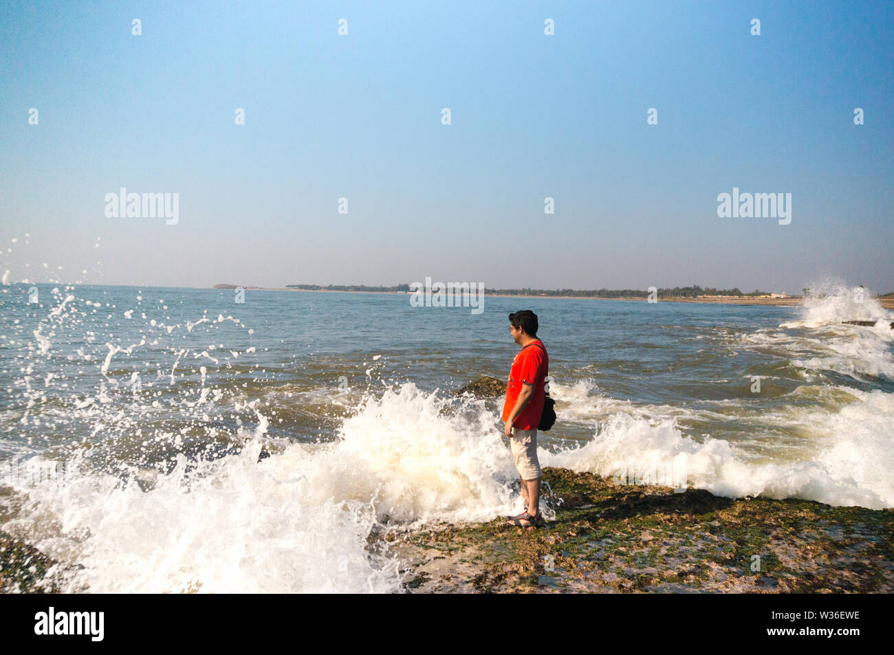 Young indian man in red standing on a rock as a wave crashes into it Stock Photo