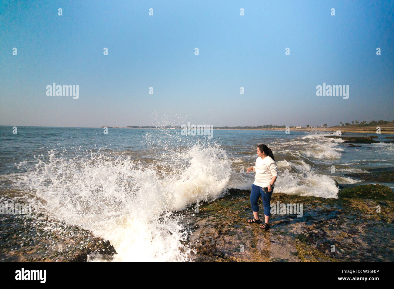 Young indian woman in white standing at a cliff as a wave splashes into it Stock Photo