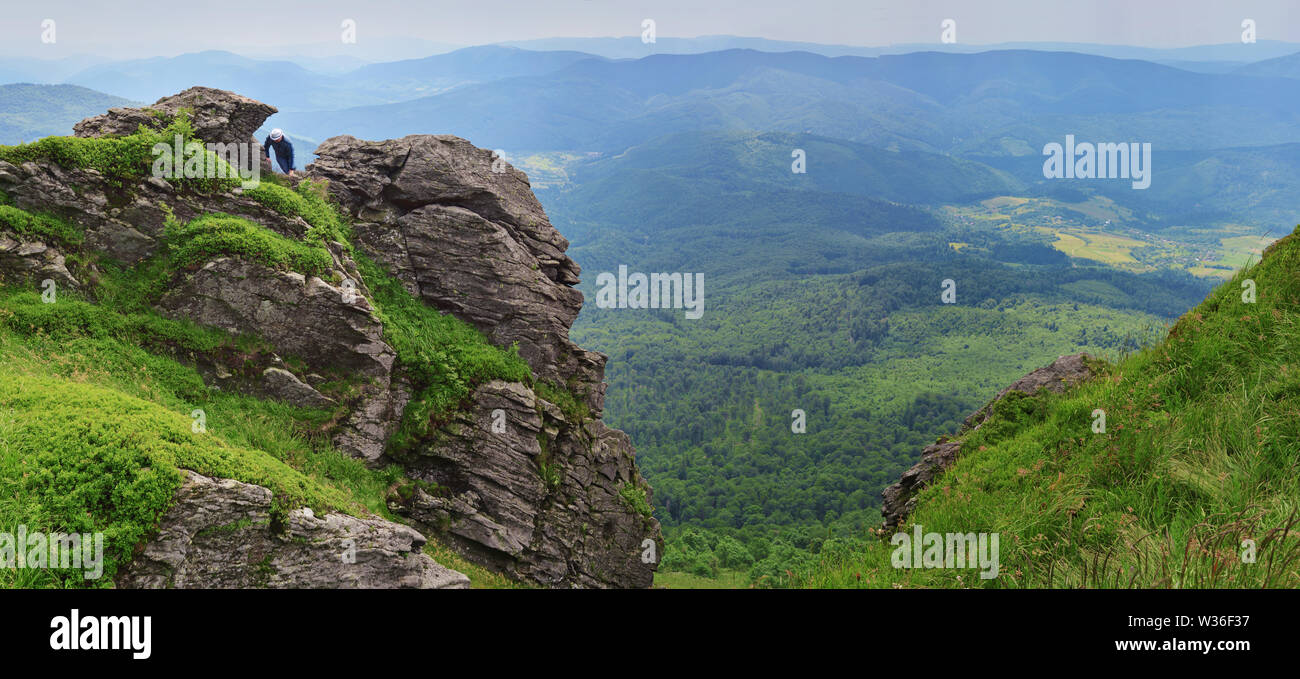 One man in a hat climbing on rocks on Pikuy mount against the valley among majestic green mountain hills covered in lush grass and forest. Cloudy summ Stock Photo