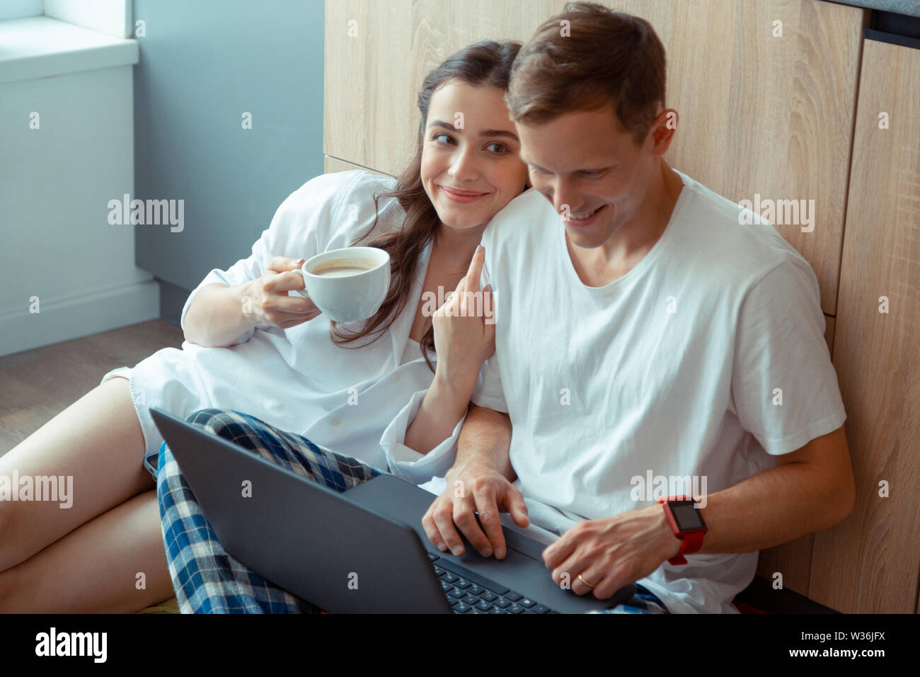 Dark-eyed wife drinking coffee while husband working on laptop Stock Photo