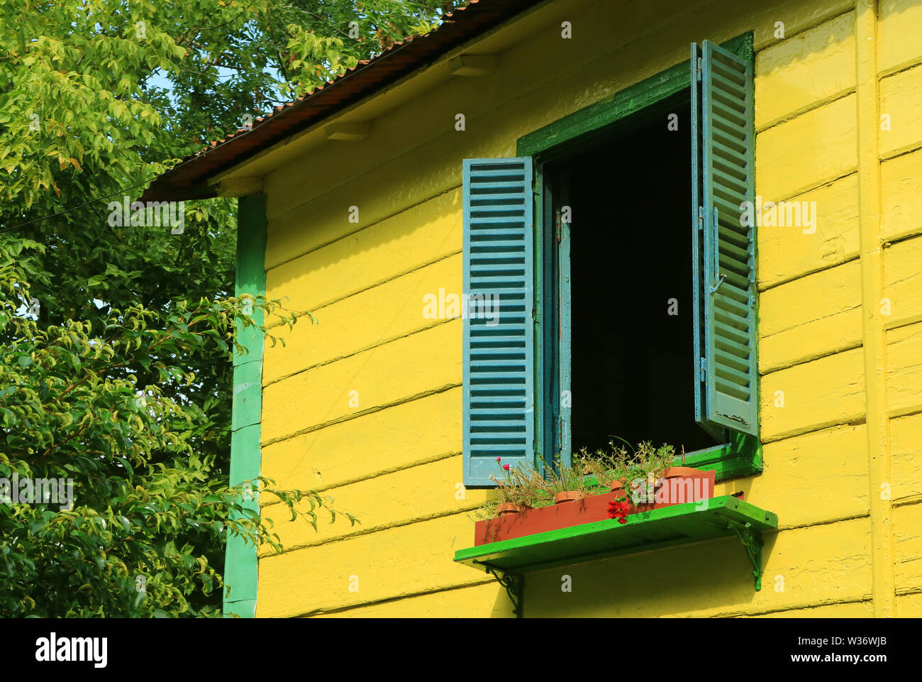 Blue wooden window shutters on vivid yellow house among green foliage in sunlight Stock Photo