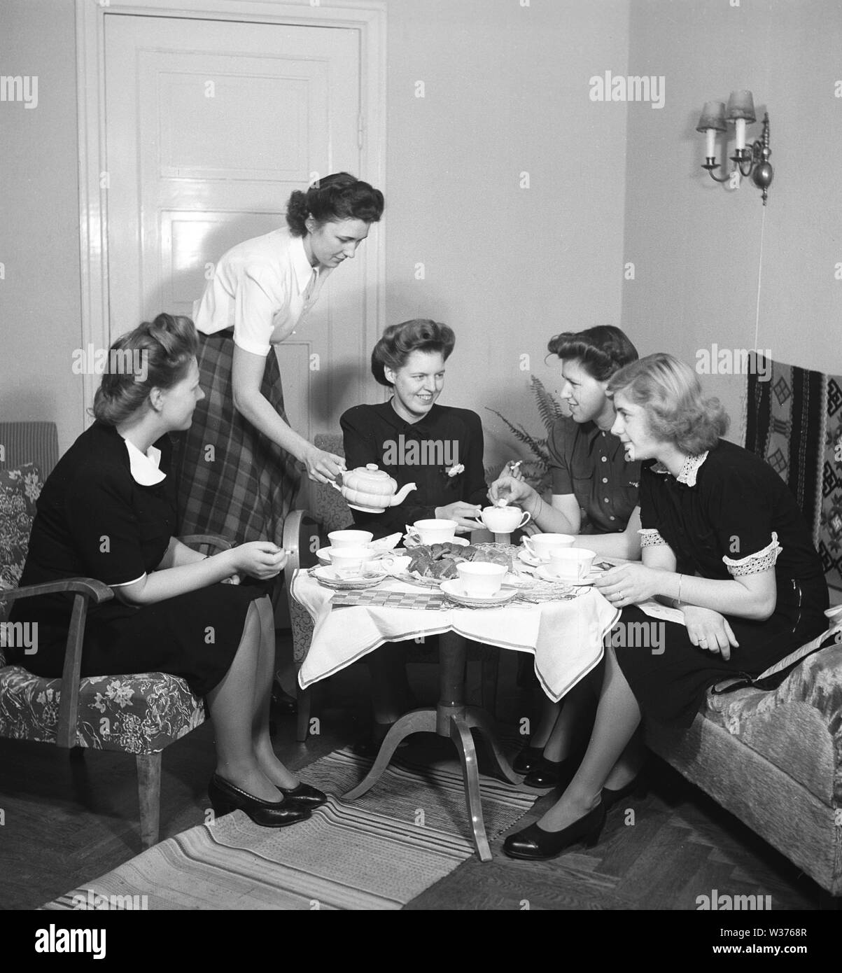 Tea time in the 1940s. A group of young women are enjoying fika themselves together around a table set with tea cups and cakes. The hostess pours the tea from a nice porcelain kettle. In the 1940s when only women met together and drank tea or coffee, was a social event. They could sit and chat, exchange gossip and news without involvement of their men or any children. Sweden 1945 Kristoffersson Ref M74-2 Stock Photo