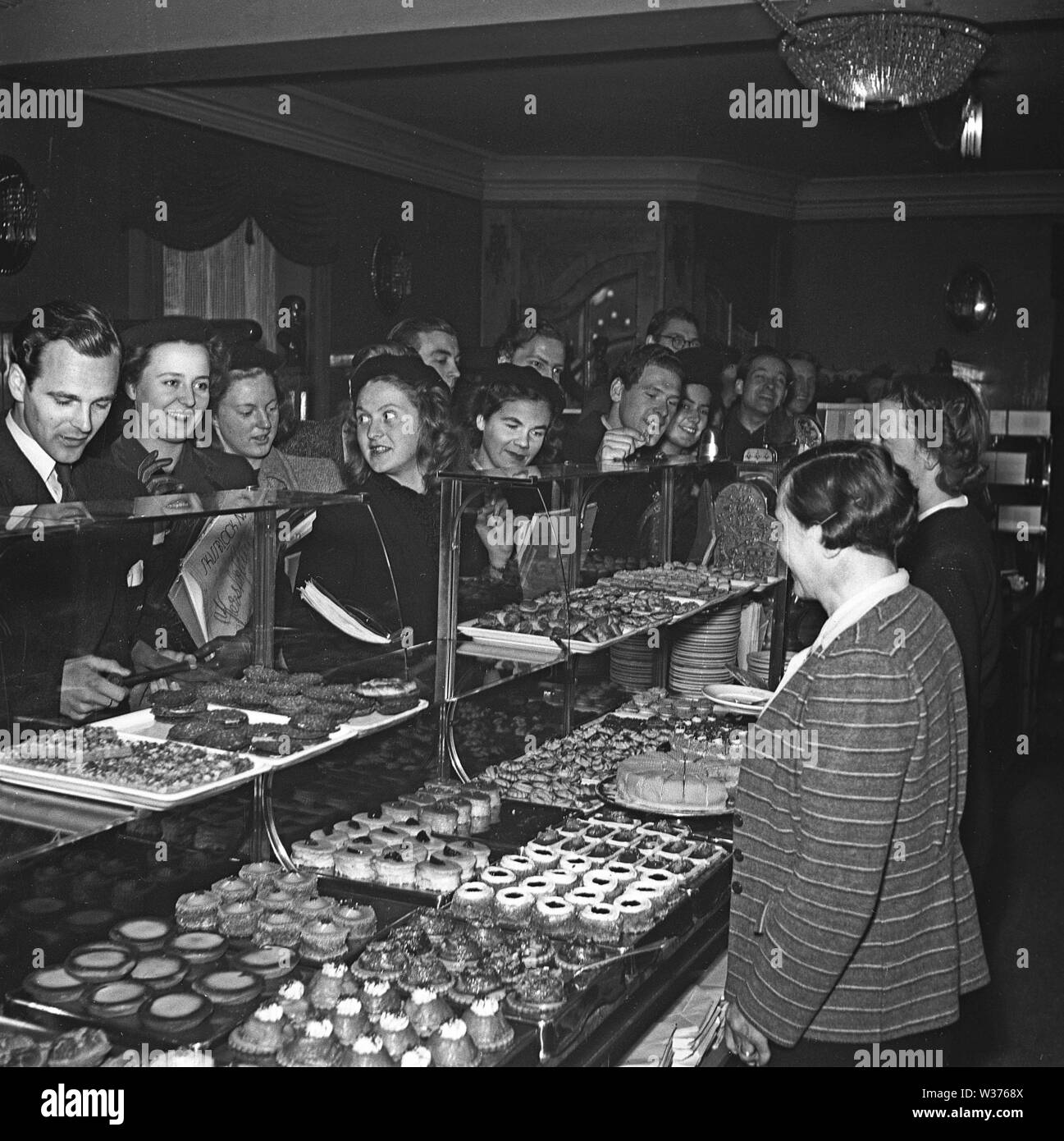 1940s coffee shop and fika time. Two women are standing behind the counter that is packed full with freshly baked cakes and pastries. A crowd of people are standing in line to order.  Sweden 1943 Kristoffersson Ref E126-2 Stock Photo