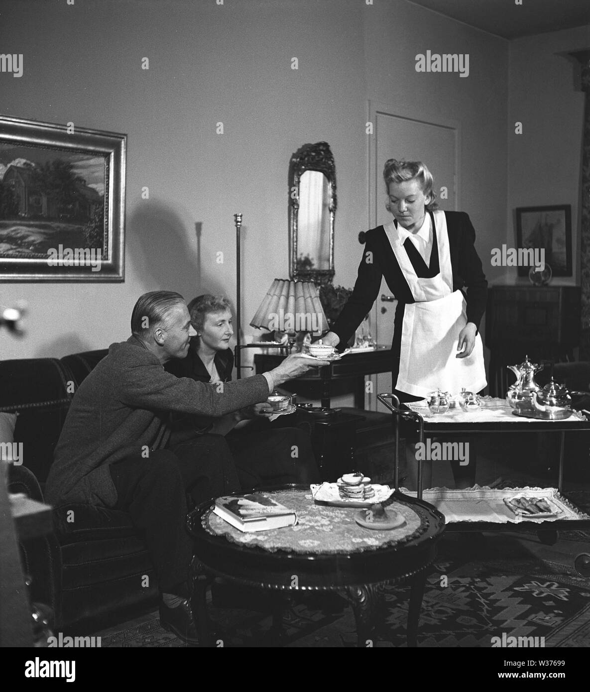 Living in the 1940s. A young woman working as a maid is serving tea and sandwiches to a man and woman sitting in the couch. She is neatly dressed in a white apron.   Sweden 1946. Kristoffersson ref X18-3 Stock Photo